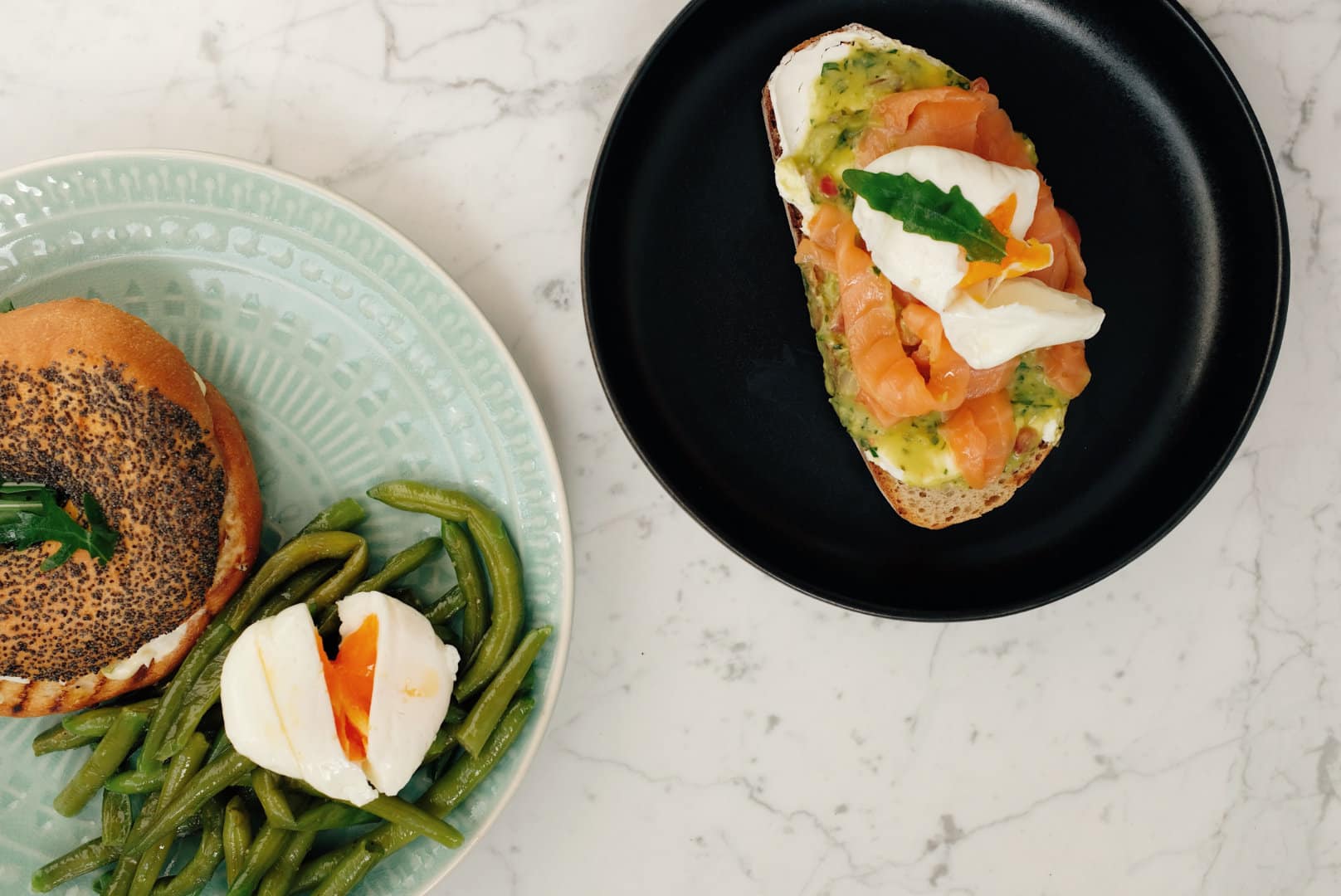 tow plates with food on them. One plate has a salmon and cream cheese bagel along with a soift boiled egg and green beans, the other a slice of toast with creme fraiche and salmon.